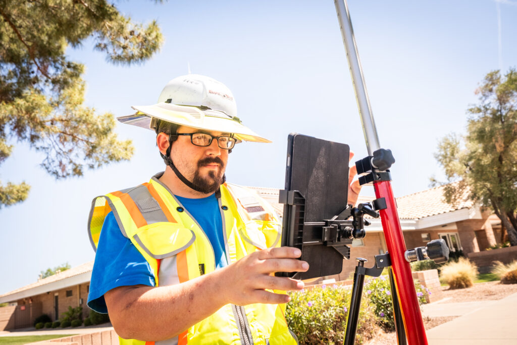 Construction worker wearing wearing a high-visibility vest with a blue t-shirt underneath looking at a tablet attached to a mapping device.