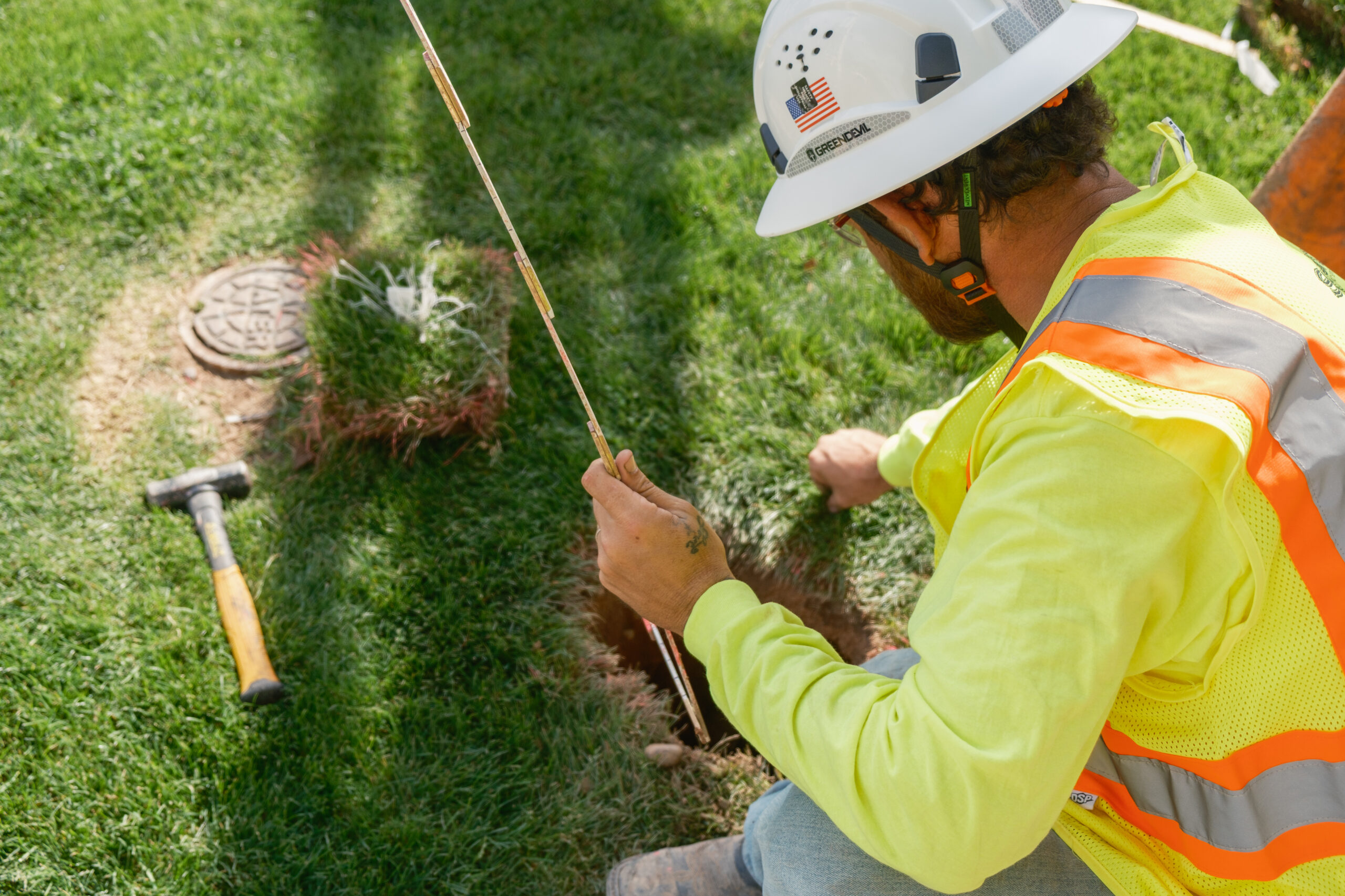 Worker in safety gear kneeling beside a small hole in the grass, feeding a rod into the ground next to a manhole cover, with a hammer lying nearby.