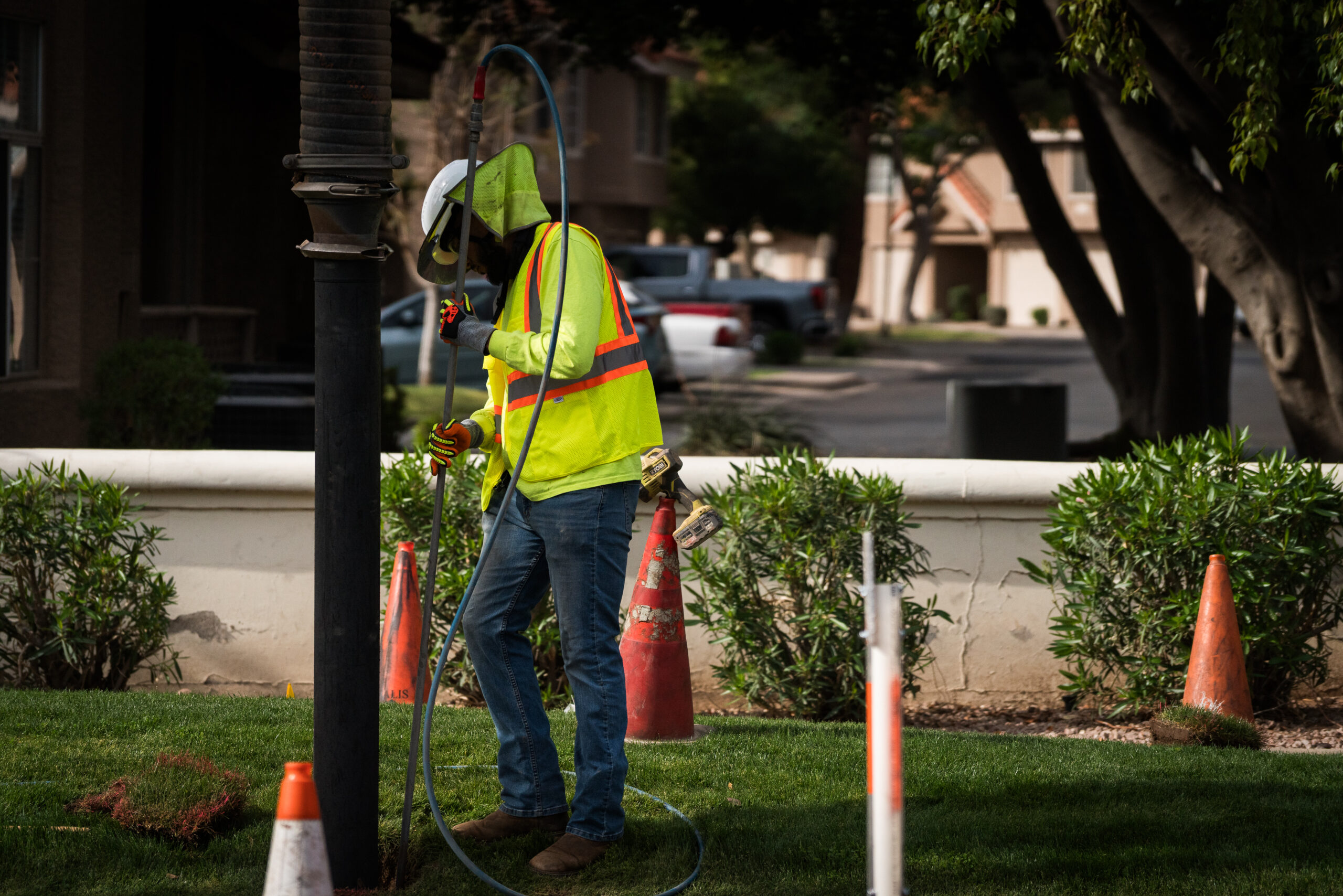 Worker in a high-visibility vest using a flexible rod tool for utility locating on a grassy area, with traffic cones nearby.