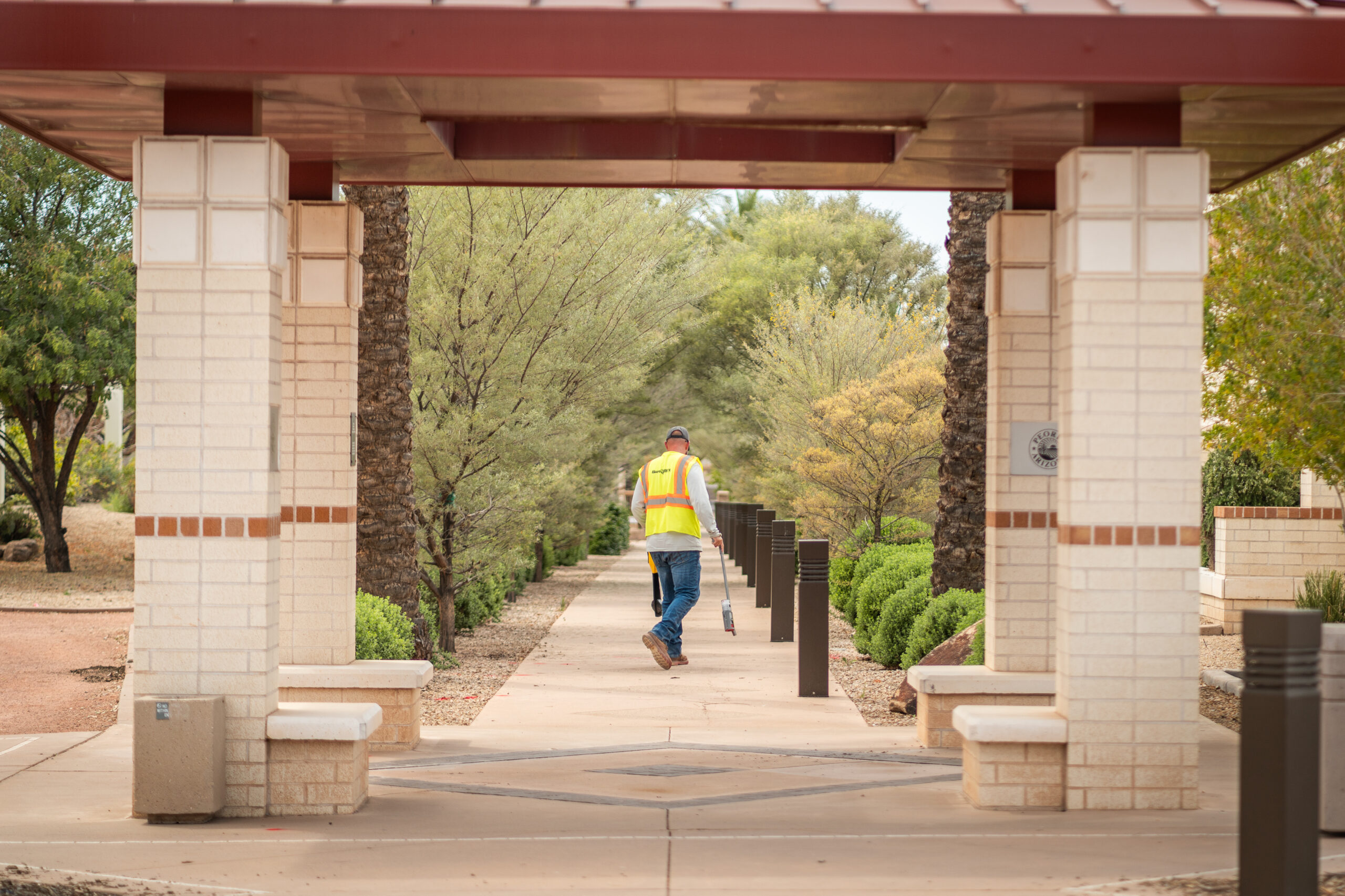 Worker in a safety vest walking along a landscaped pathway, holding utility locating equipment.