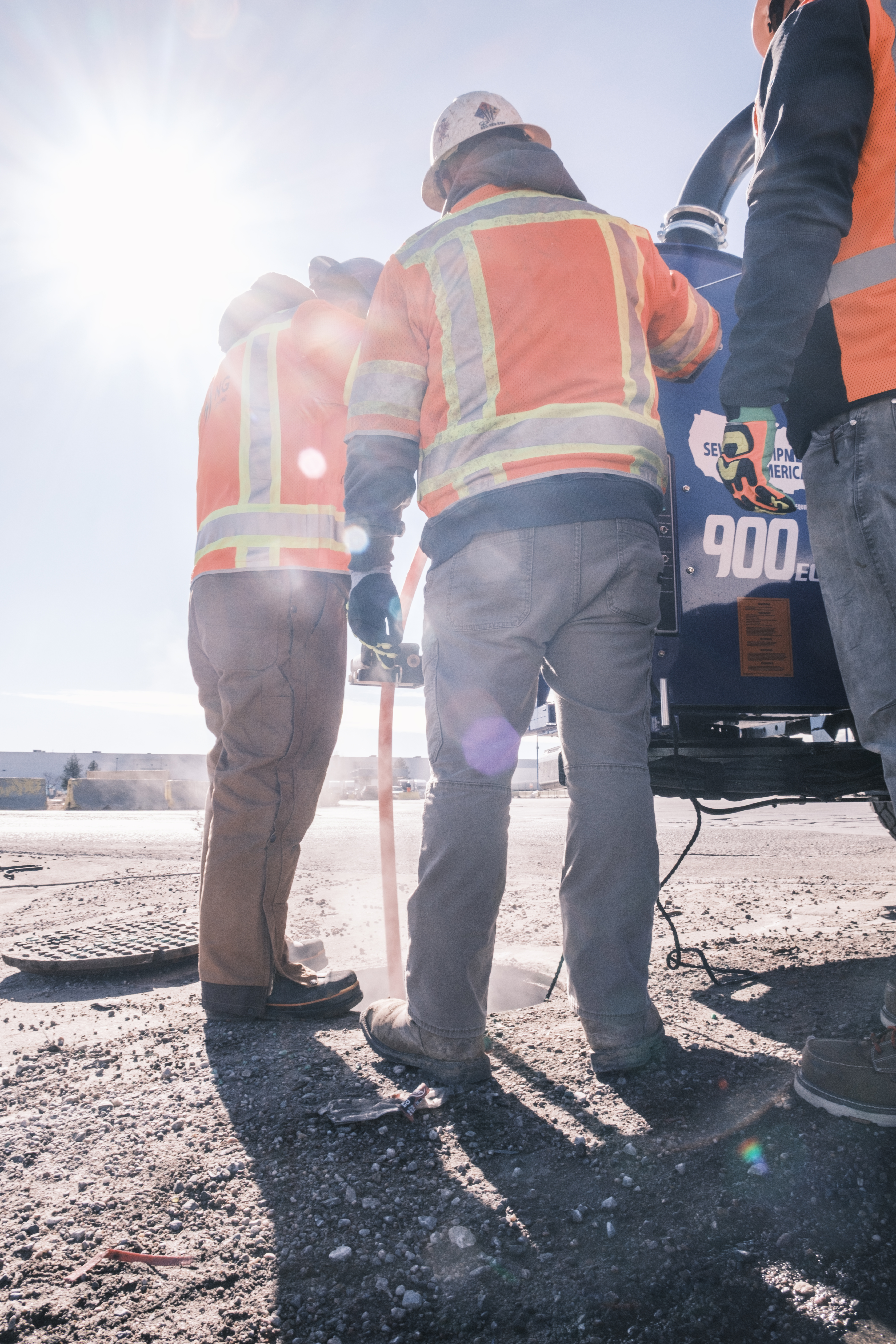 Close-up of two workers in orange safety vests and hard hats operating vacuum excavation equipment, with the sun shining brightly behind them.