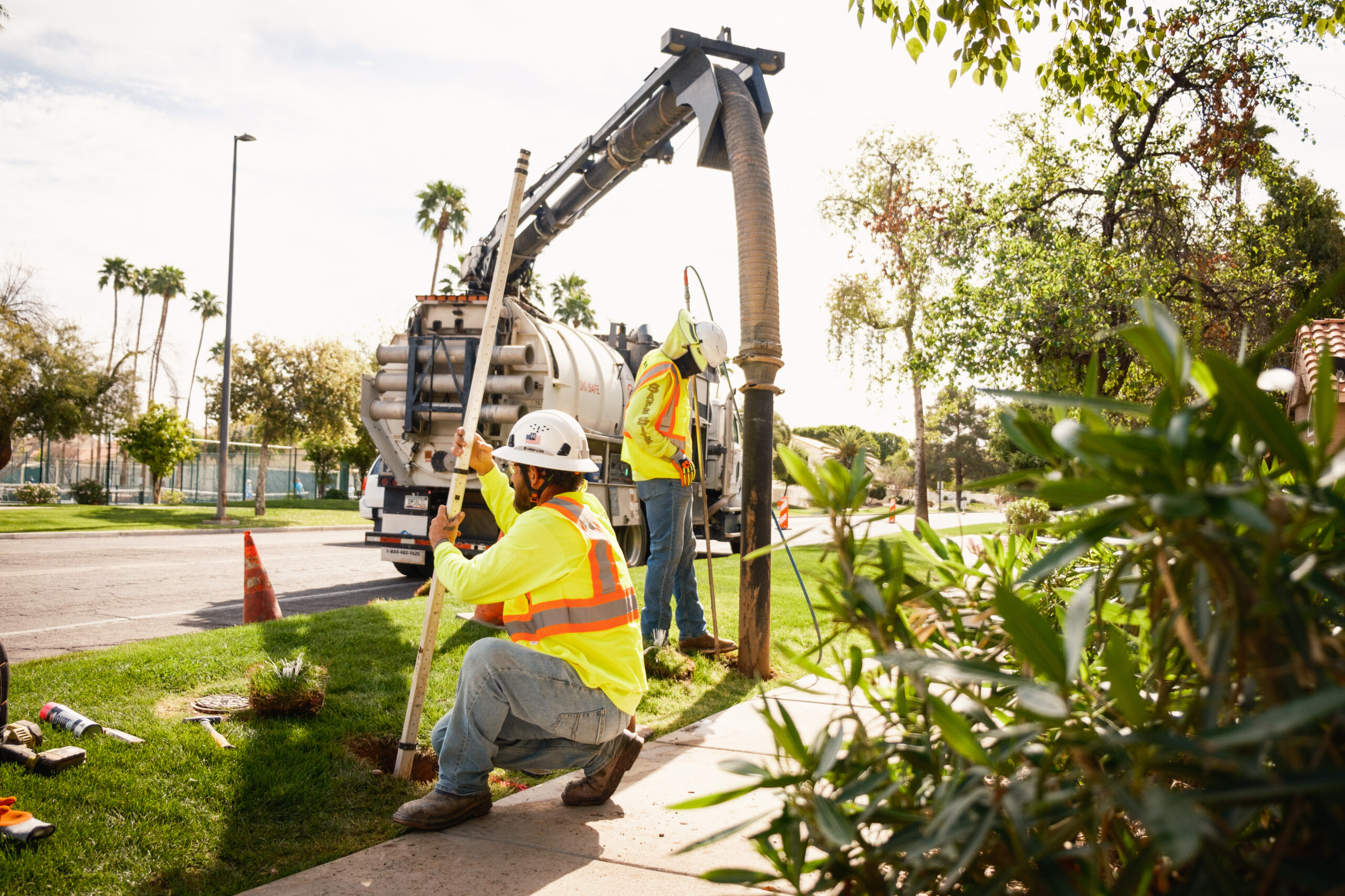 Two construction workers in high-visibility vests and hard hats utilizing excavation equipment on a vehicle to dig a whole in a grass patch next to a sidewalk.
