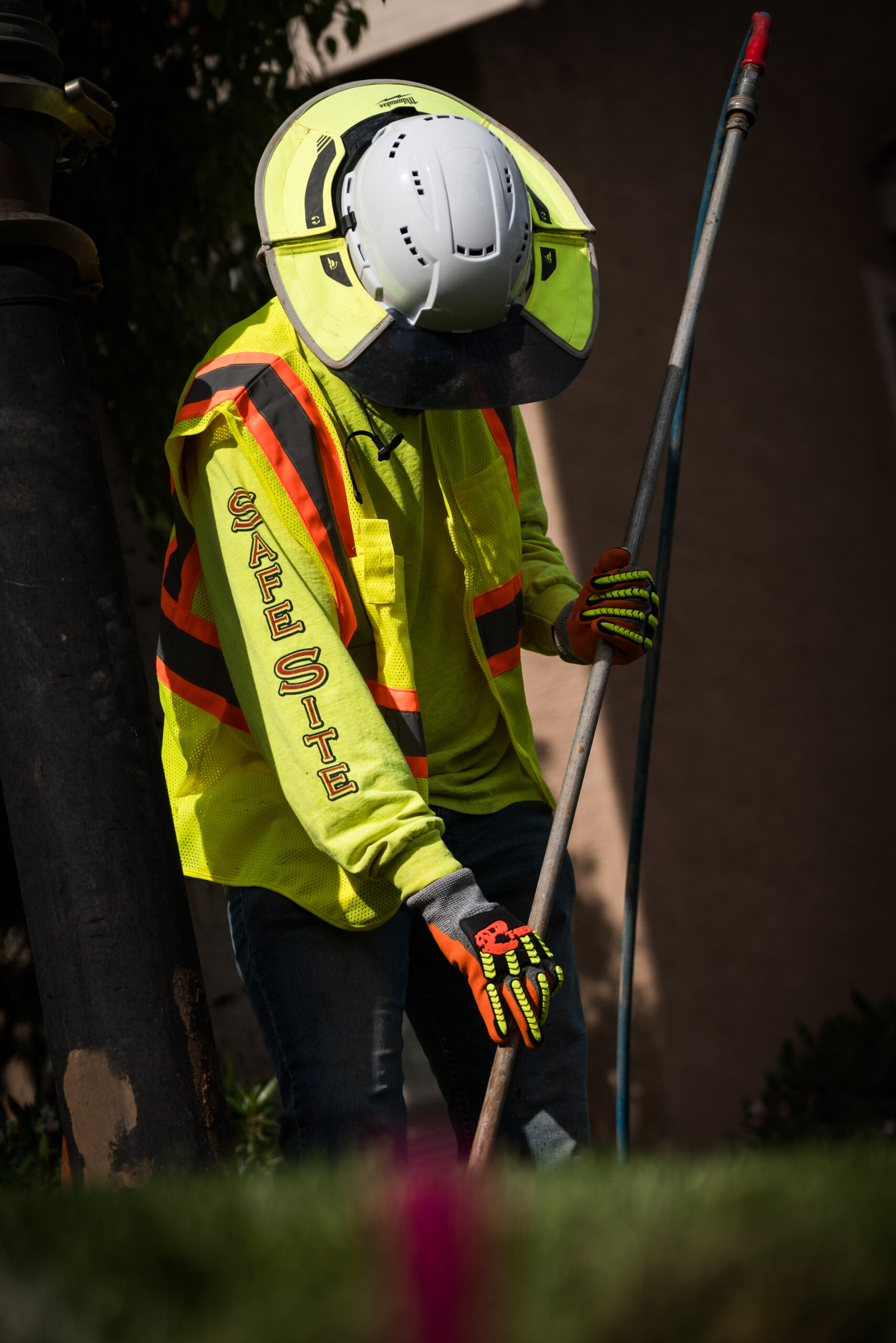 Construction worker wearing a high-visibility vest and a sweater underneath with safesite vertical down the sleeve. He has excavation equipment in his hand while looking towards the ground.