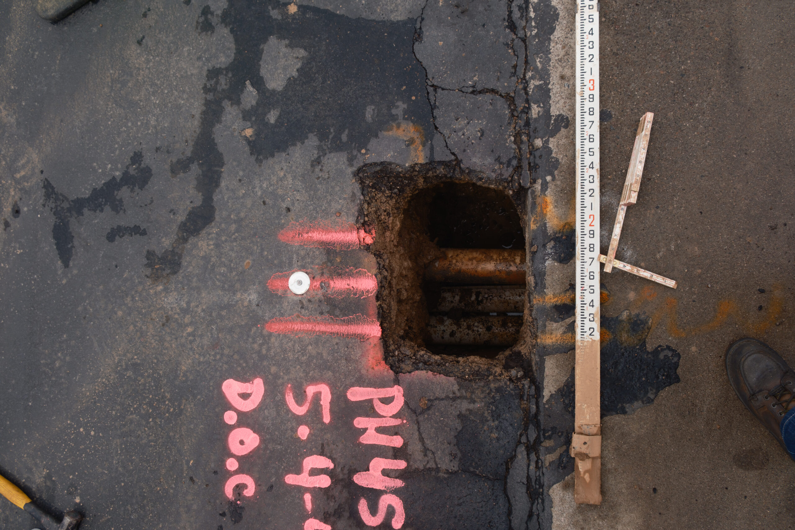 Hole dug up on street road exposing pipes underground, writing is visible on the road pointing to the hole.