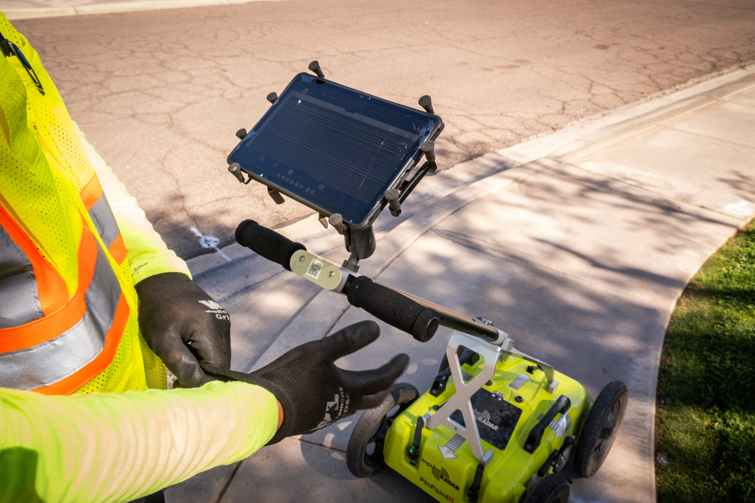 Safe Site-220 A close-up of a person operating a yellow ground-penetrating radar device with a mounted tablet, standing on a sidewalk.