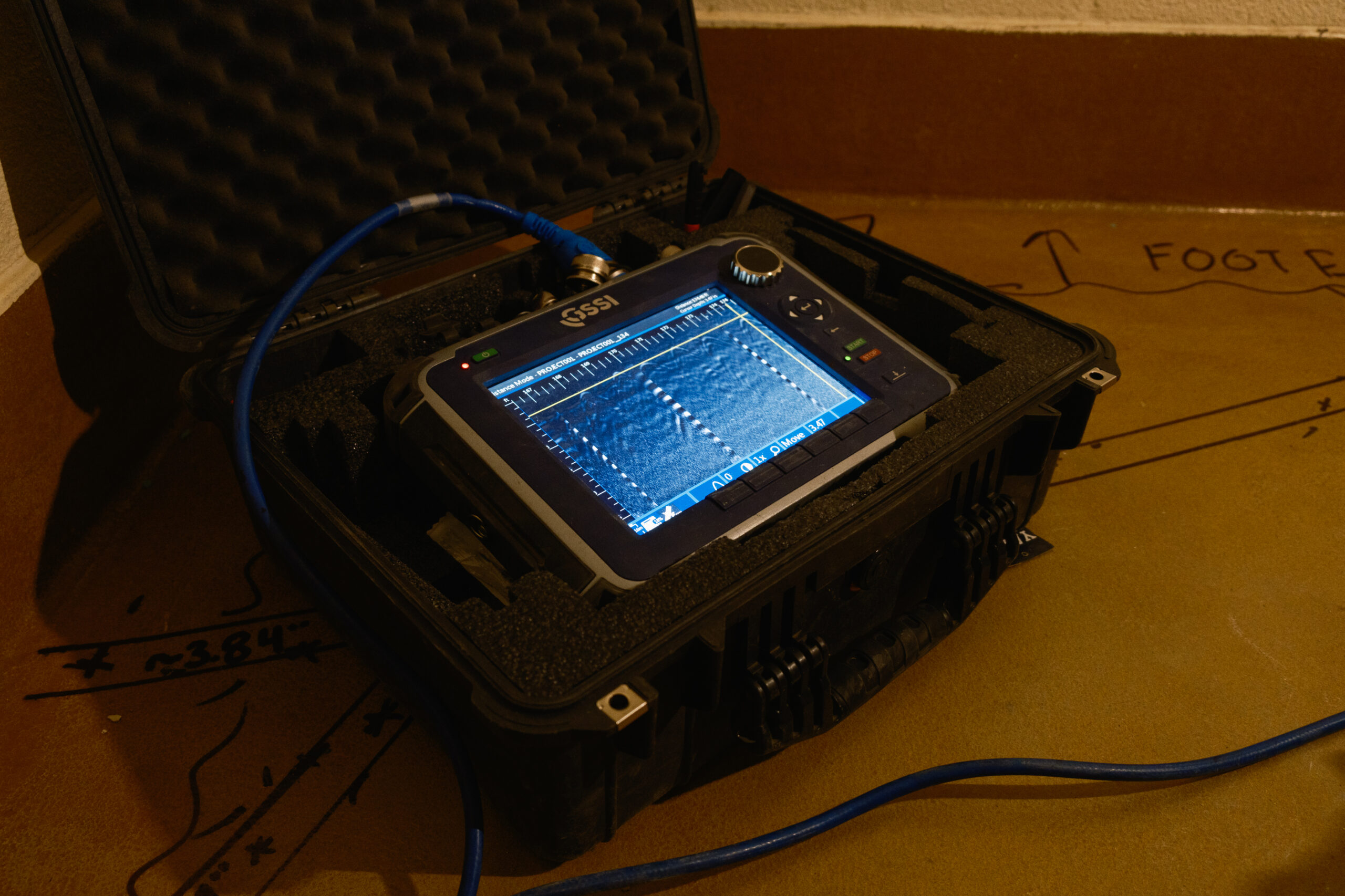 Close-up of a GSSI ground-penetrating radar control unit in a protective case, displaying scan data on its screen.