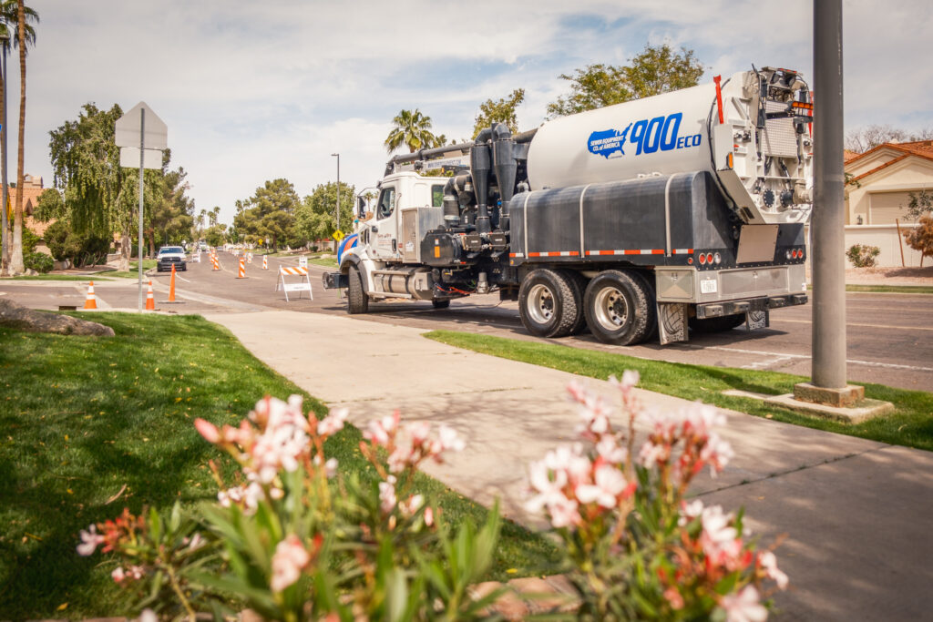 A vacuum excavation truck parked on a residential street with traffic cones set up, viewed from behind flowers in the foreground.