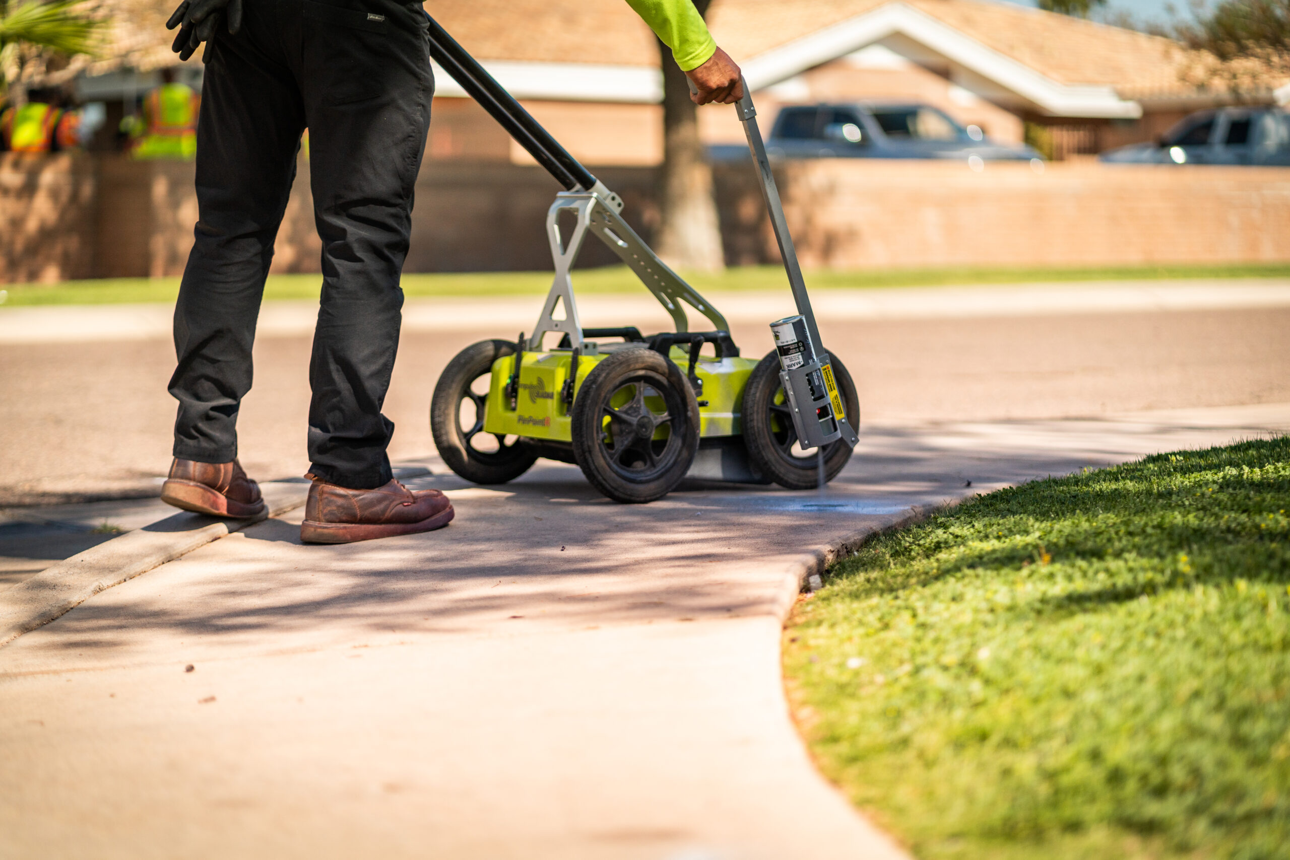 Side view of a ground-penetrating radar device on a sidewalk, with an operator standing nearby wearing dark pants and brown shoes.