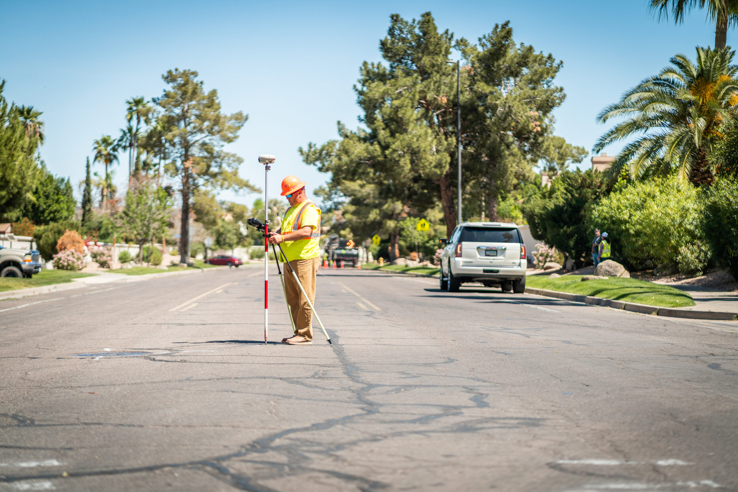 A construction worker in a high-visibility vest and hard hat stands in the middle of a residential street using surveying equipment, surrounded by trees, parked cars, and houses.